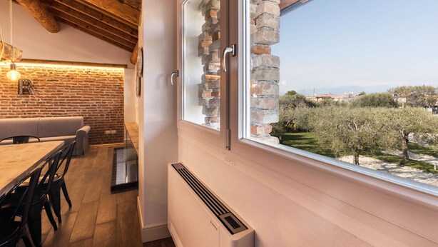 A room with a brick wall, a wooden ceiling, a dining table, and a window with a view of trees and buildings. - Large Family Apartment "Rovere" with Wi-Fi, Shared Garden & Pool (Desenzano)