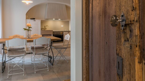 A dining area with a wooden table and chairs, a hanging light fixture, and a view into a kitchen with a stove and oven. - Apartment "Ortensia" in Country House Style with Private Garden & Shared Pool (Desenzano)