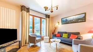 A modern living room with a flat-screen TV, a grey sofa, a white coffee table, and a grey armchair. There is a large window with curtains, a painting on the wall, and a chandelier. - Coët A Tous by Interhome (Carnac)