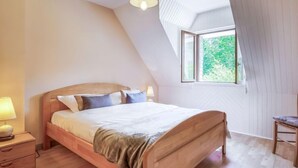 A bedroom with a sloped ceiling, a bed with a wooden headboard, a bedside table with a lamp, and a window with a view of greenery. - Coët A Tous by Interhome (Carnac)