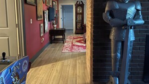 A hallway with a wooden floor, a red wall with framed pictures, a grandfather clock, and a knight statue. - Harry Potter Castle (Greer)