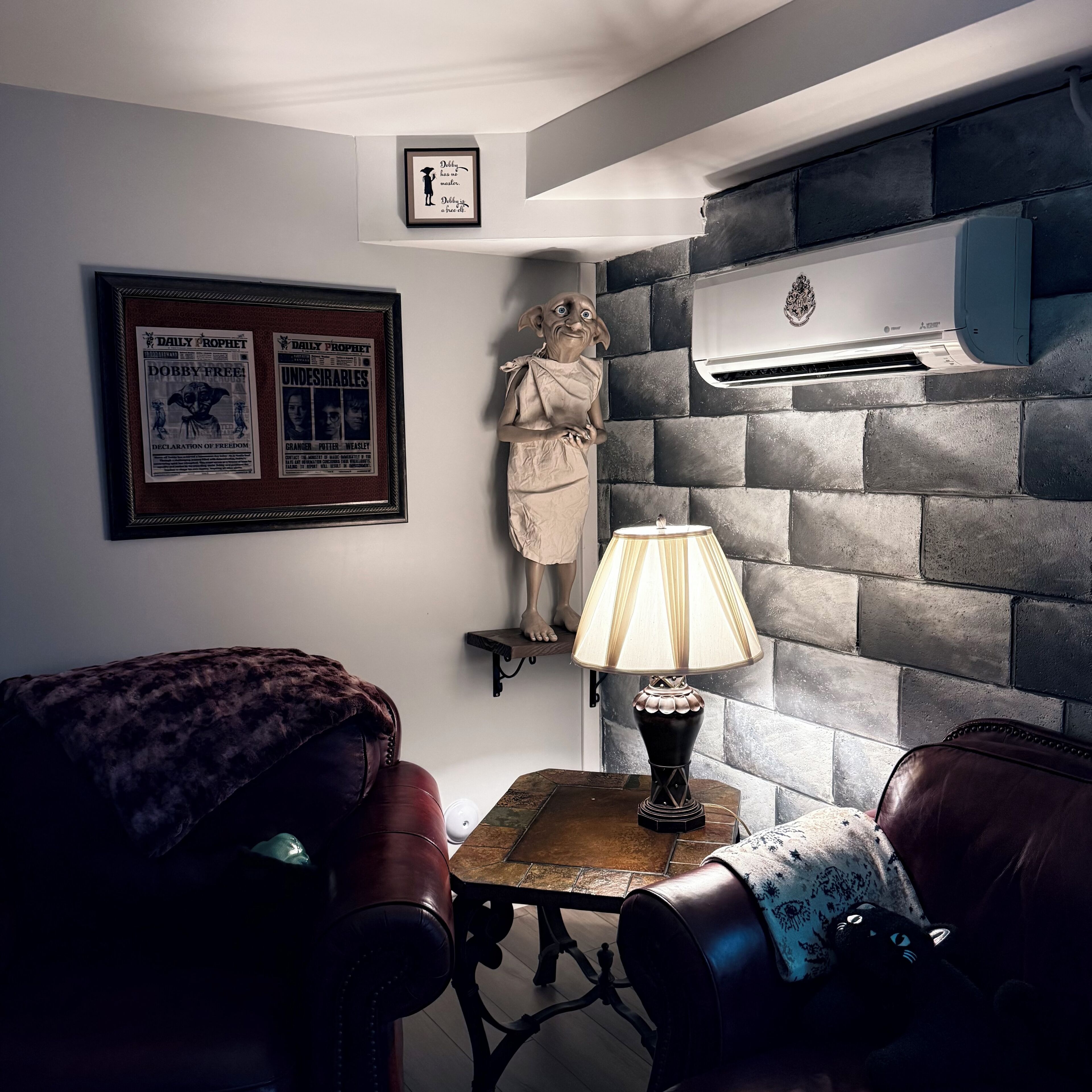 A room with a stone wall, a framed poster, a statue, a table with a lamp, and leather chairs.