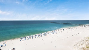 A beach with white sand, a pier extending into the water, and numerous umbrellas.