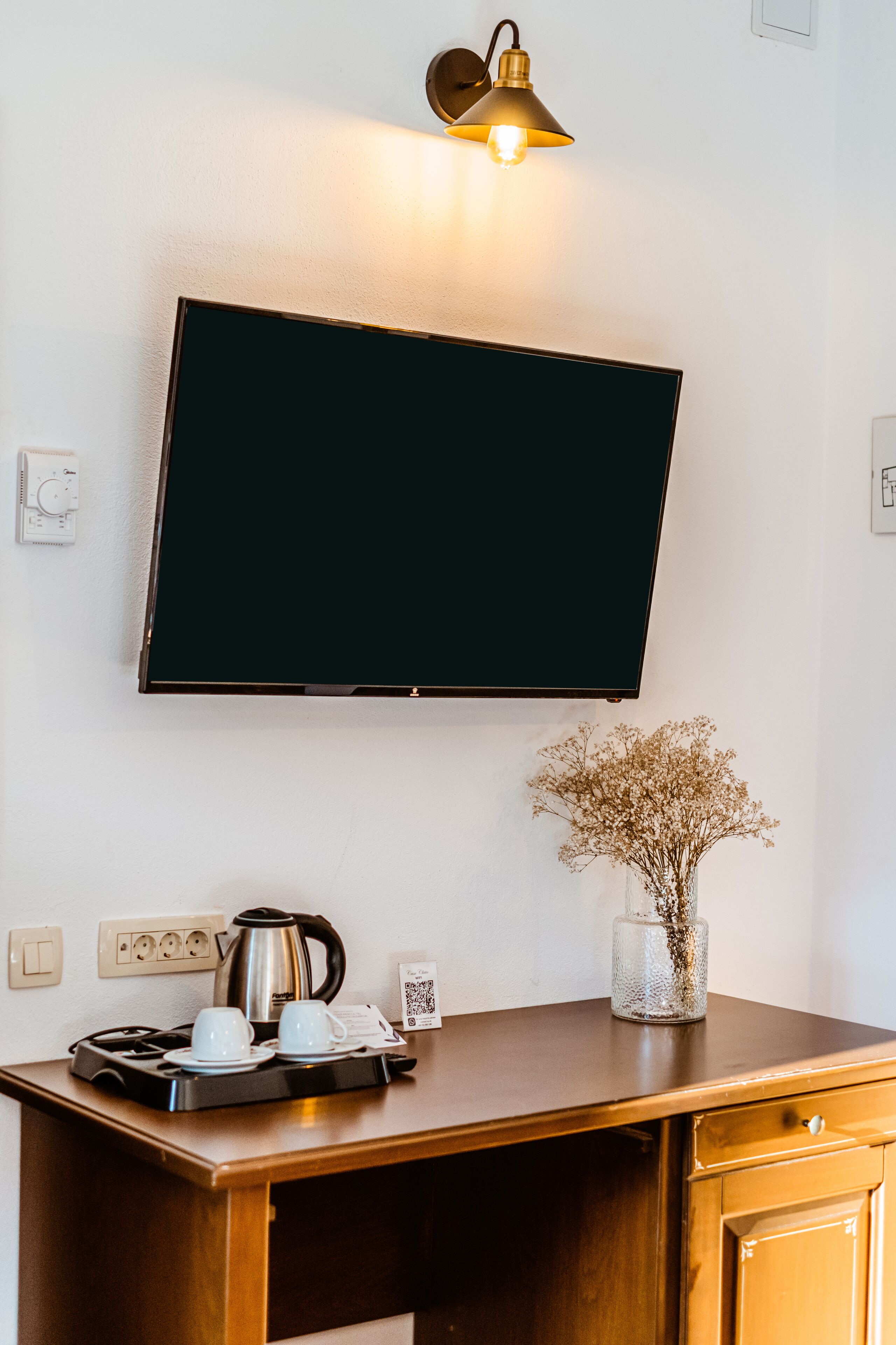Une pièce équipée d’un téléviseur à écran plat fixé au mur, d’une machine à café, de tasses, d’un vase contenant des fleurs séchées et d’un bureau en bois.