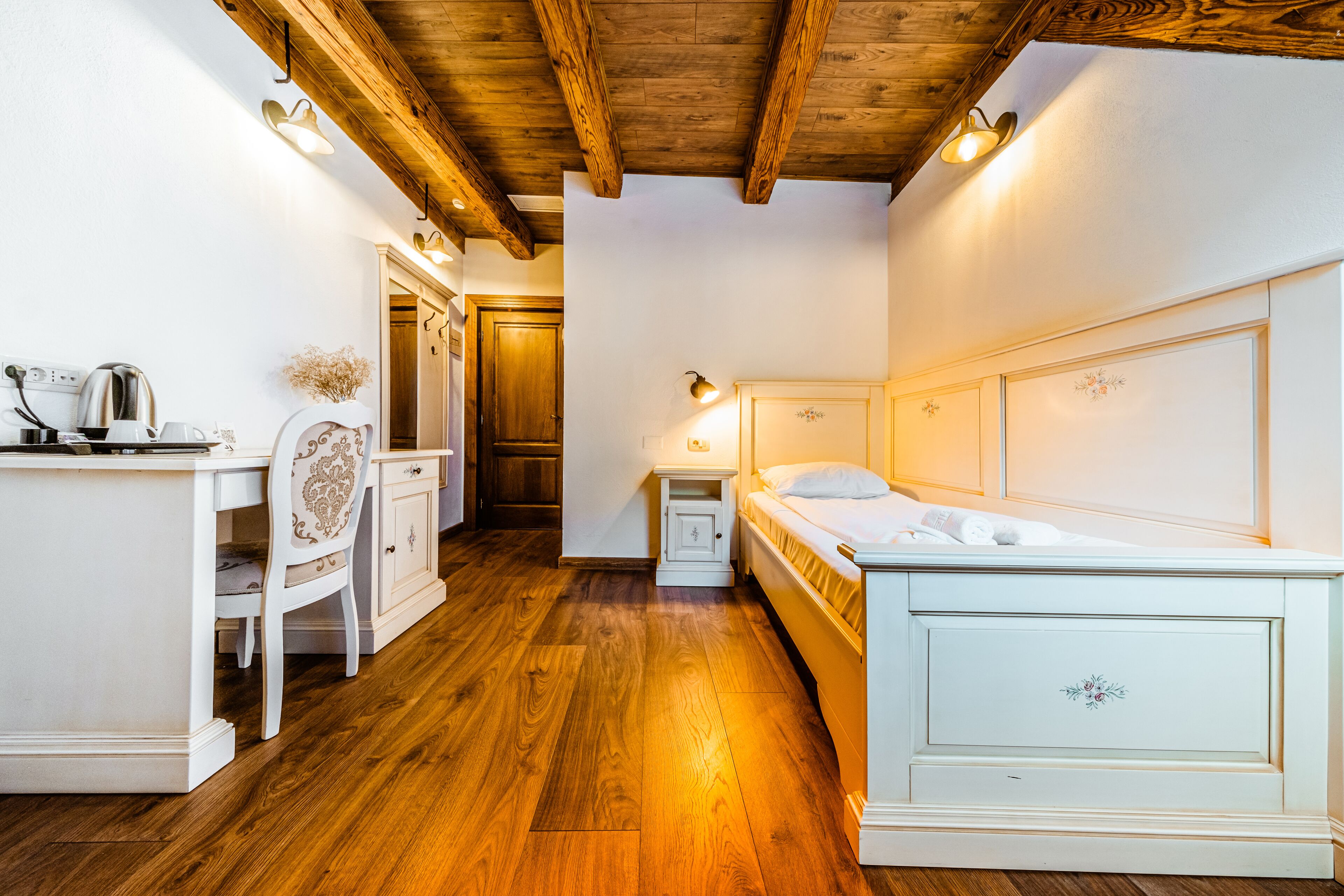 A room with a wooden floor, a bed with a headboard, a desk with a chair, a nightstand, and a wall-mounted light fixture.