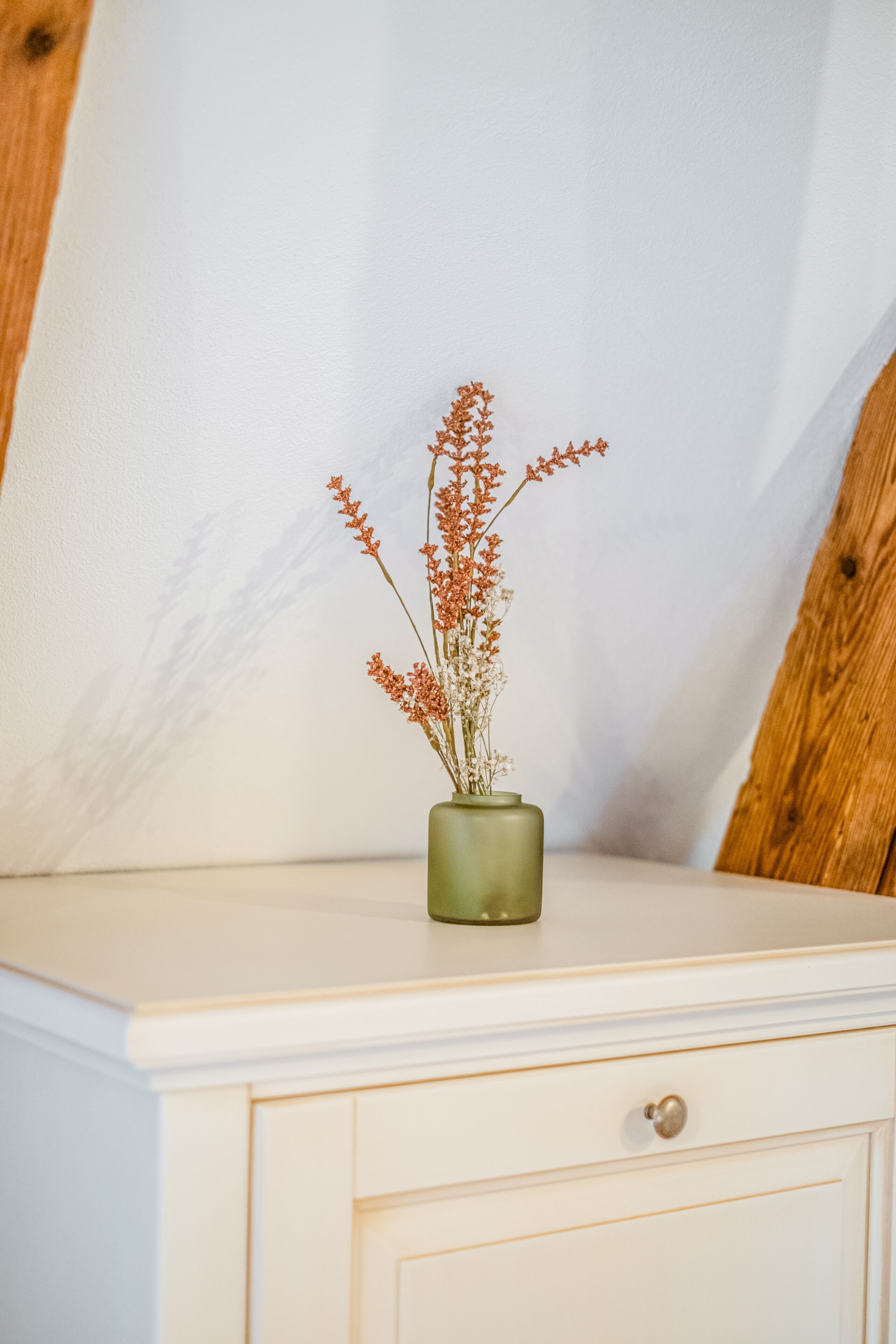 A green vase with dried flowers on a white bedside table against a plain wall.