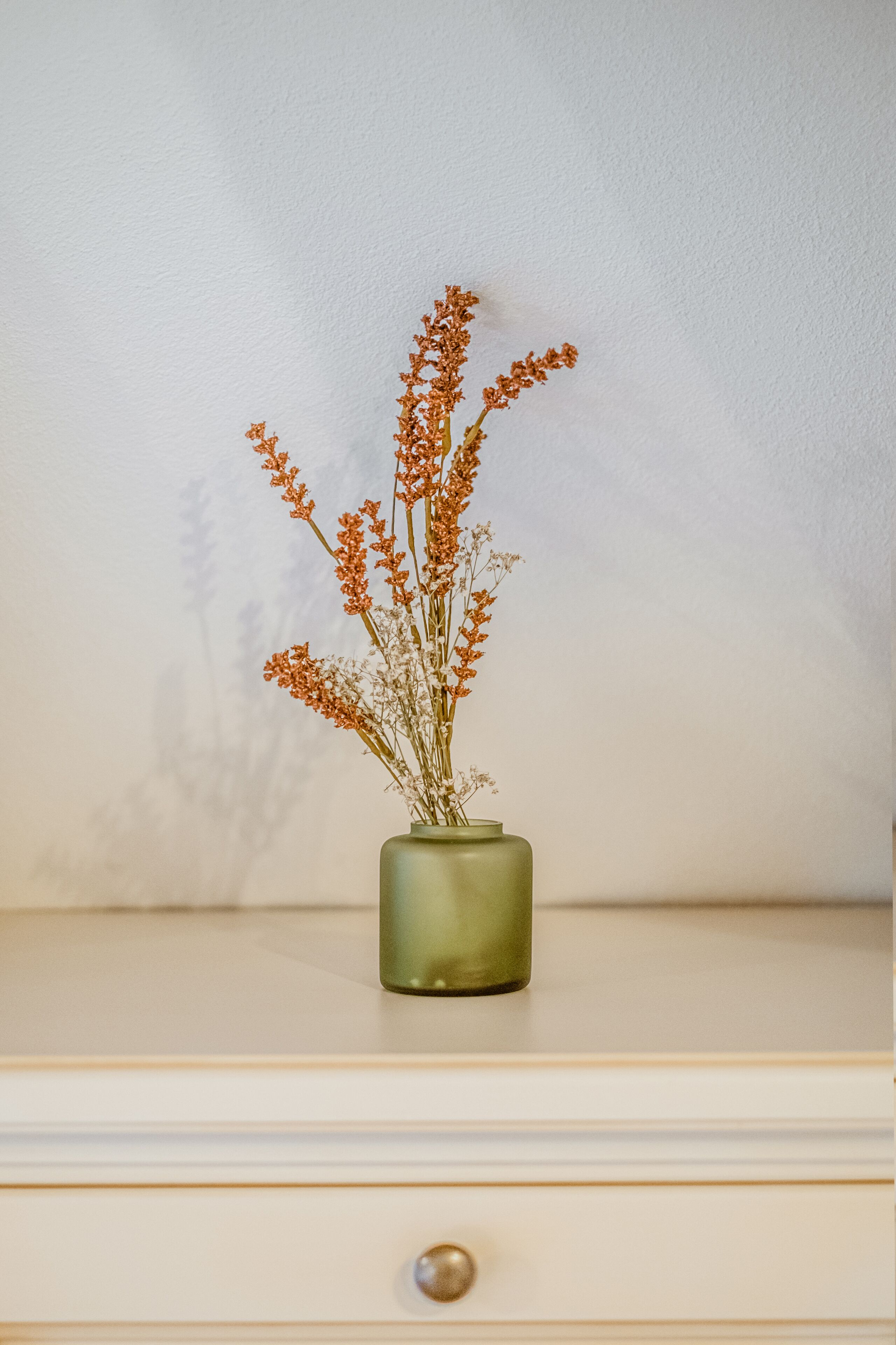 Eine Vase mit getrockneten Blumen auf einer Kommode vor einer schlichten Wand.