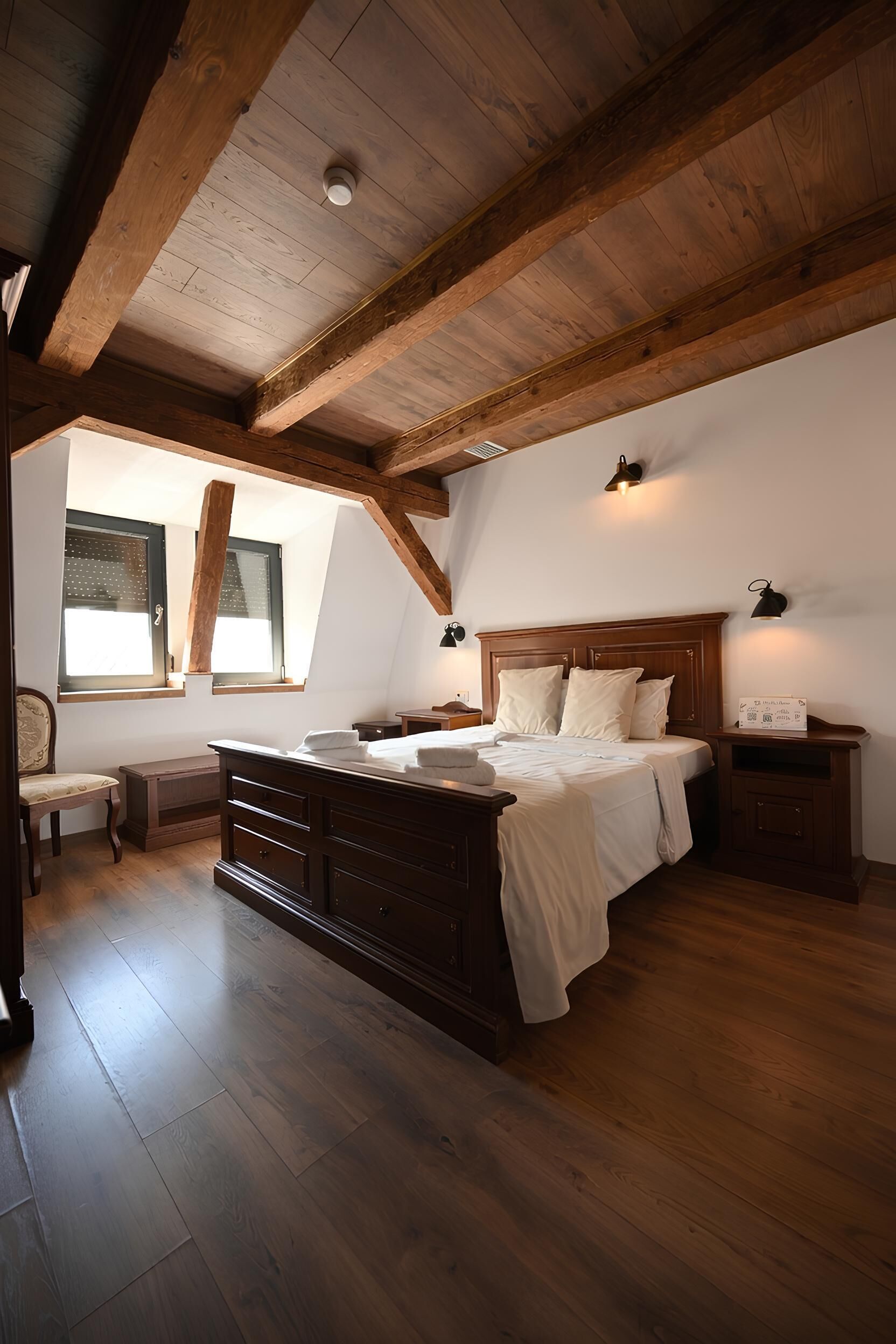 Un dormitorio con techo de madera, cama con ropa de cama blanca, mesita de noche, una silla y una ventana con persianas.
