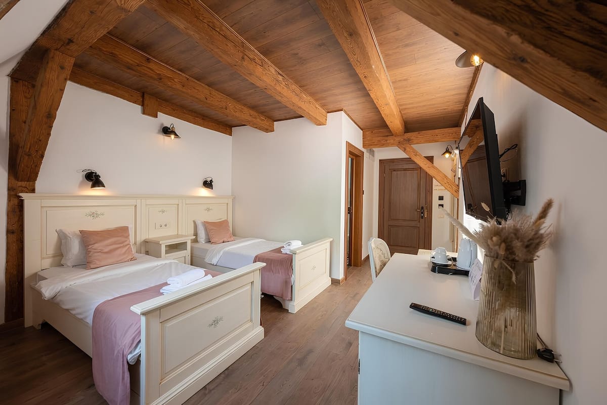 A bedroom with two beds, wooden beams, a desk, a chair, a TV, and a vase with dried flowers.
