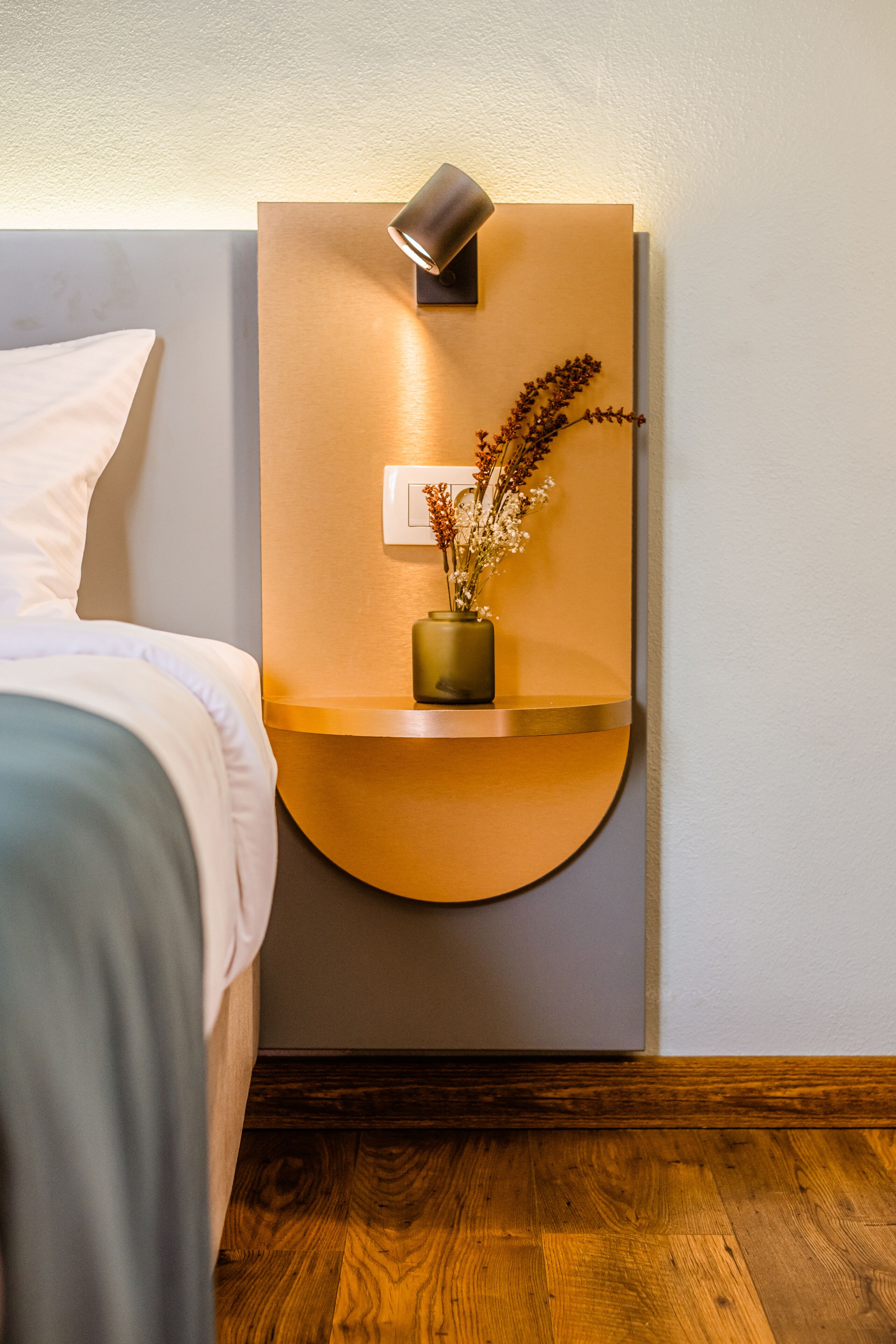 Ein modernes Schlafzimmer mit einem Bett, einer Wandablage mit einer Vase getrockneter Blumen und einer Nachttischlampe.