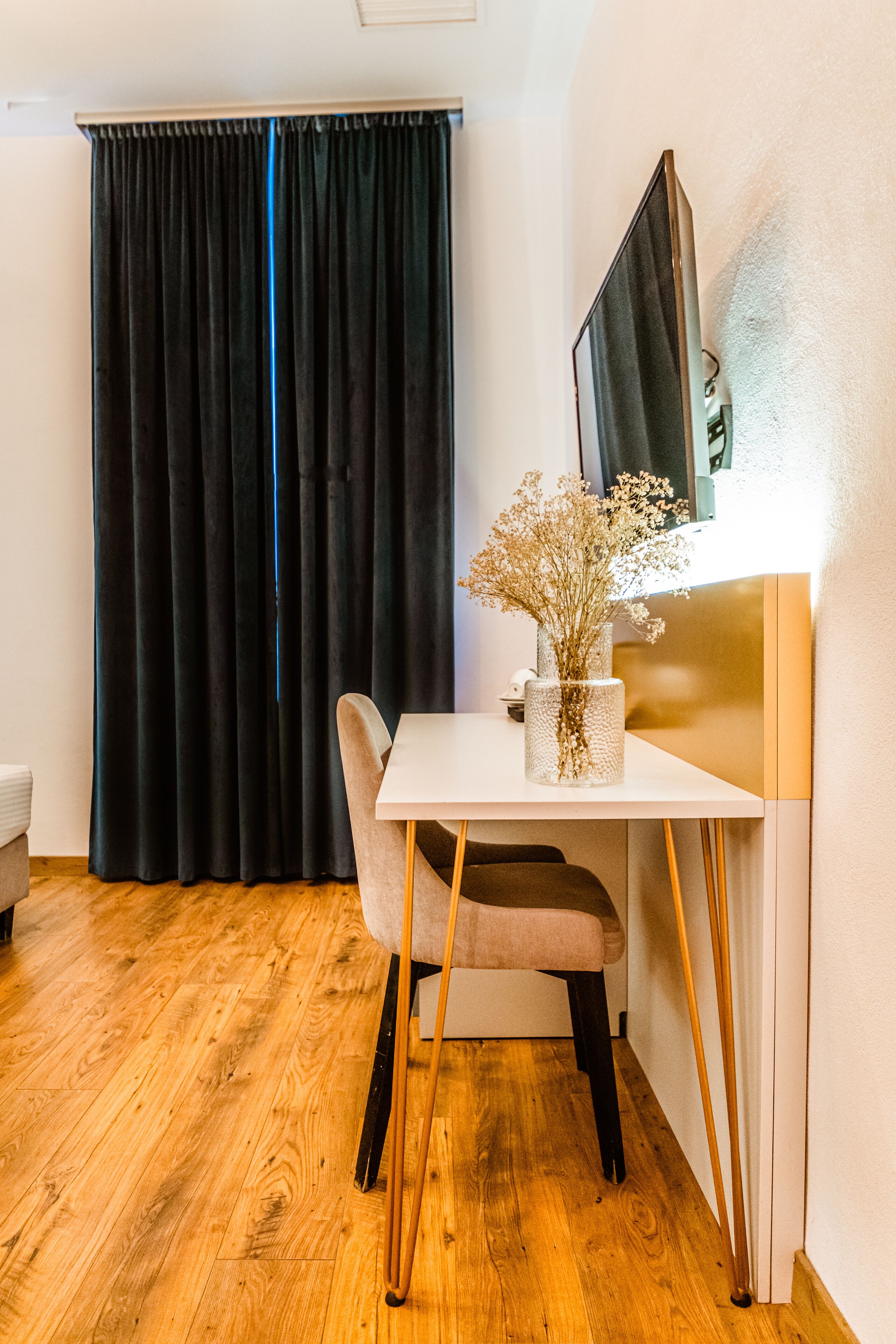 A room with a white desk, a chair, a vase with dried flowers, a black curtain, and a wooden floor.