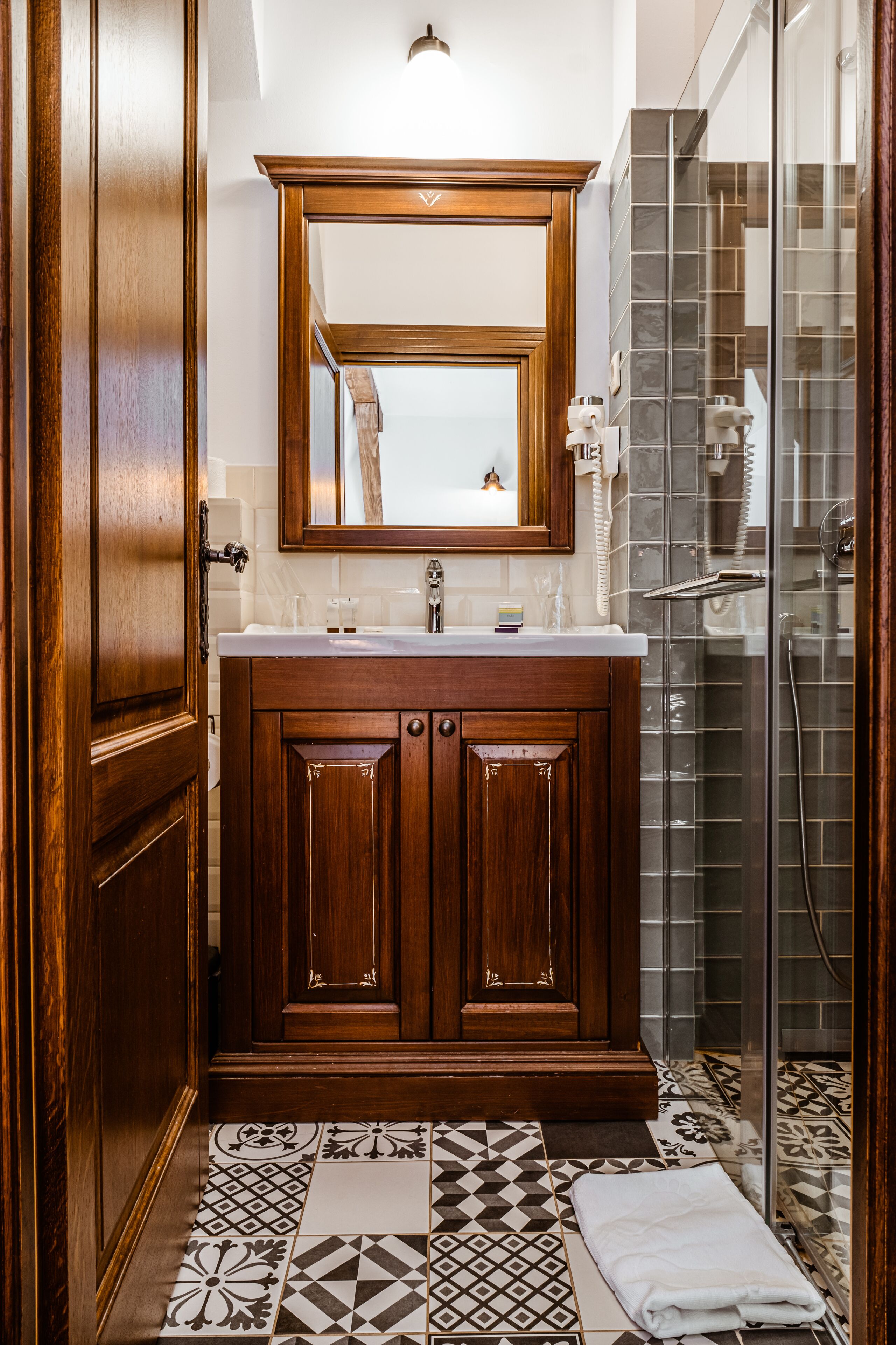A bathroom with a wooden vanity, a large mirror, a shower with glass doors, and patterned floor tiles.