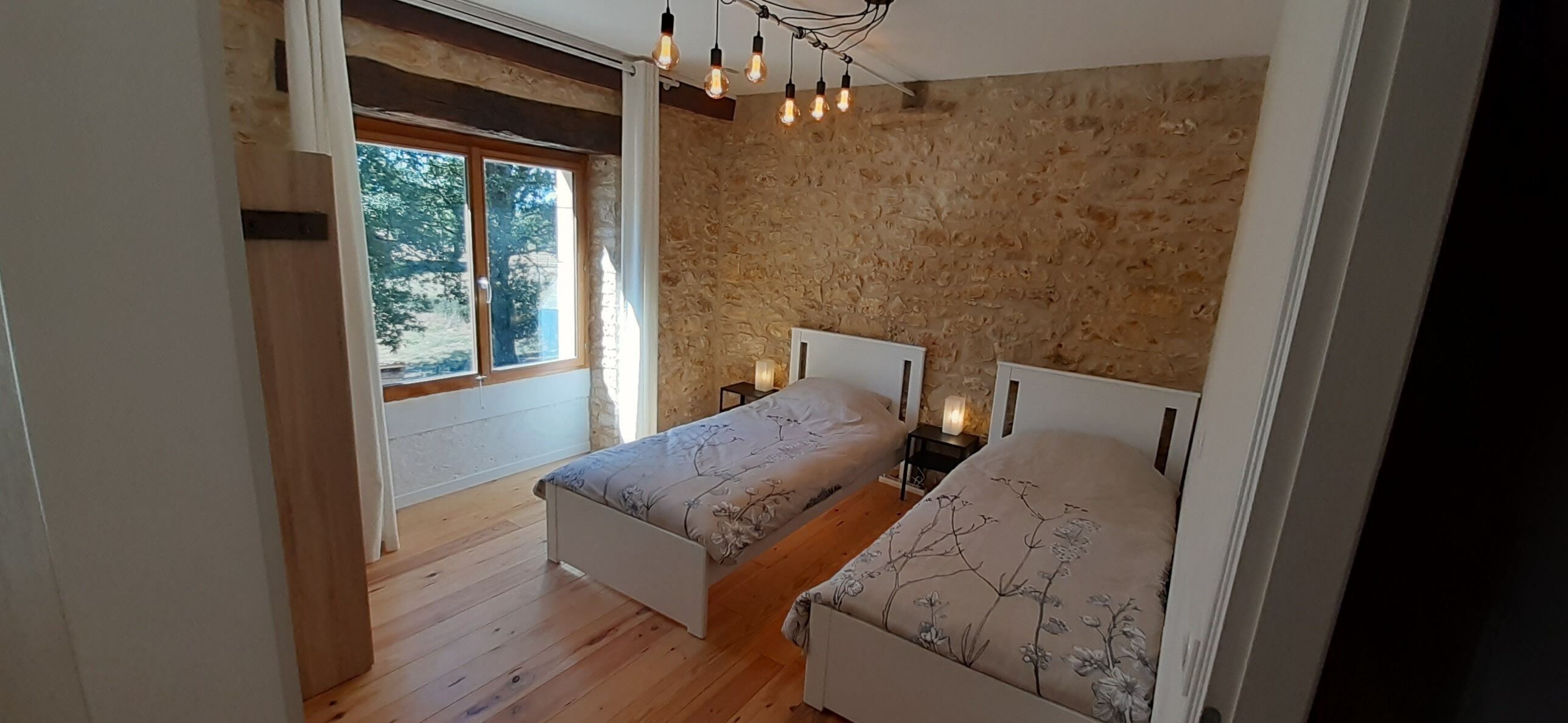 Une chambre avec un plancher en bois, un mur en pierre, deux lits avec une literie à motifs floraux, une table de chevet avec une lampe et une fenêtre avec des rideaux.