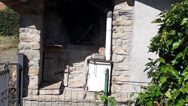 Un edificio in pietra con un camino, un'area recintata con un bidone e un albero di fronte.