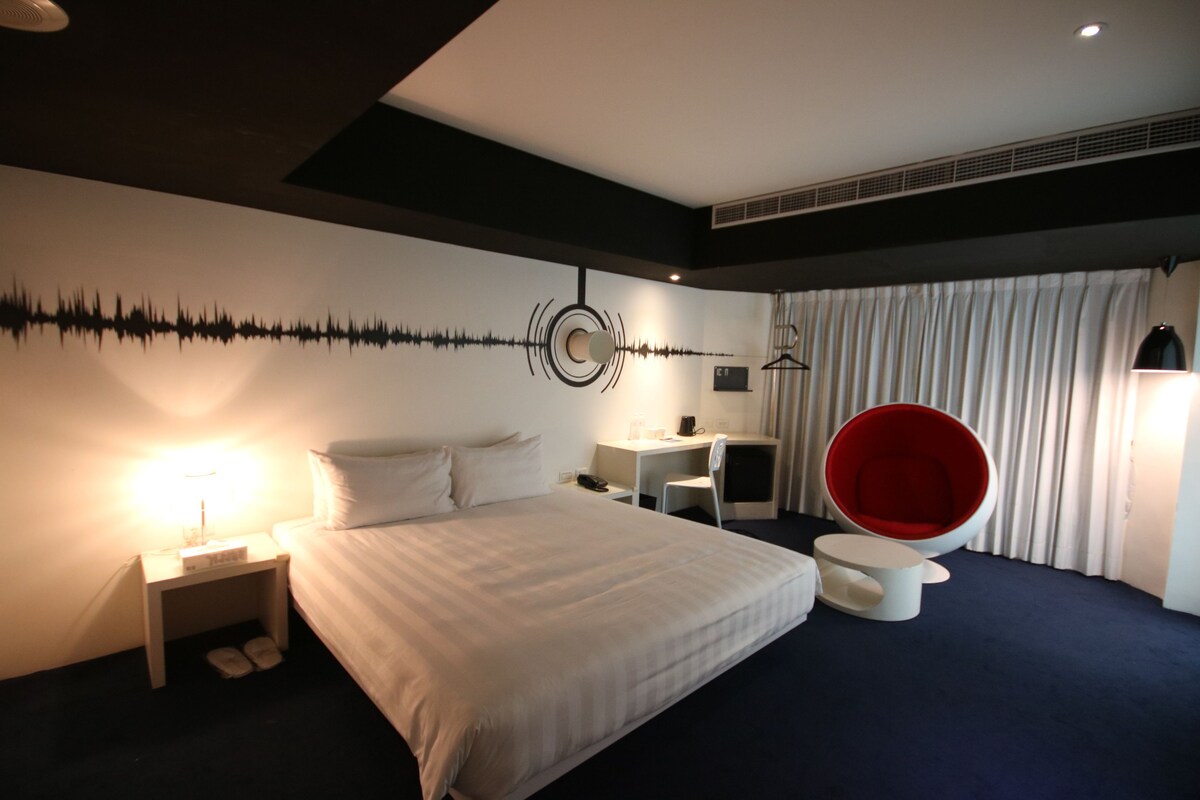 Habitación de hotel con una cama grande, un escritorio, una silla roja y una pared con diseño de onda sonora.