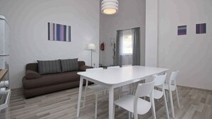 Uma sala de estar moderna com um sofá castanho, uma mesa branca e cadeiras. Há um abajur, um extintor de incêndio e arte abstrata nas paredes.