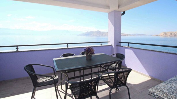 Varanda com mesa e cadeiras, com vista para um corpo de água e montanhas ao longe.