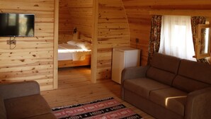 A cozy cabin interior with a wooden ceiling, a flat-screen TV mounted on the wall, a sofa, a patterned rug, and a bed in the background. - Kazdagları Saglikli Yasam Tatil Koyu (Edremit)