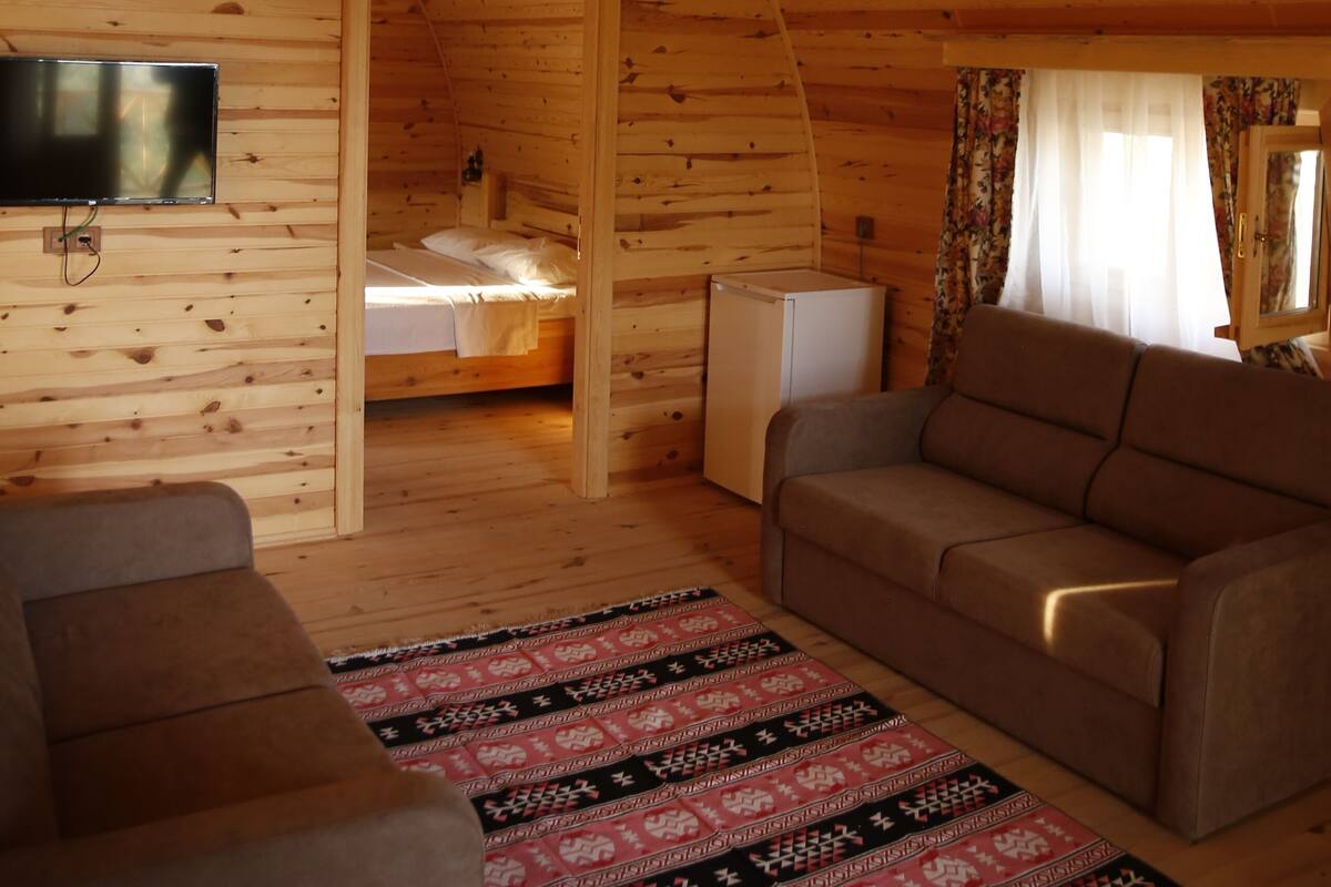 A cozy cabin interior with a wooden ceiling, a flat-screen TV mounted on the wall, a sofa, a patterned rug, and a bed in the background.