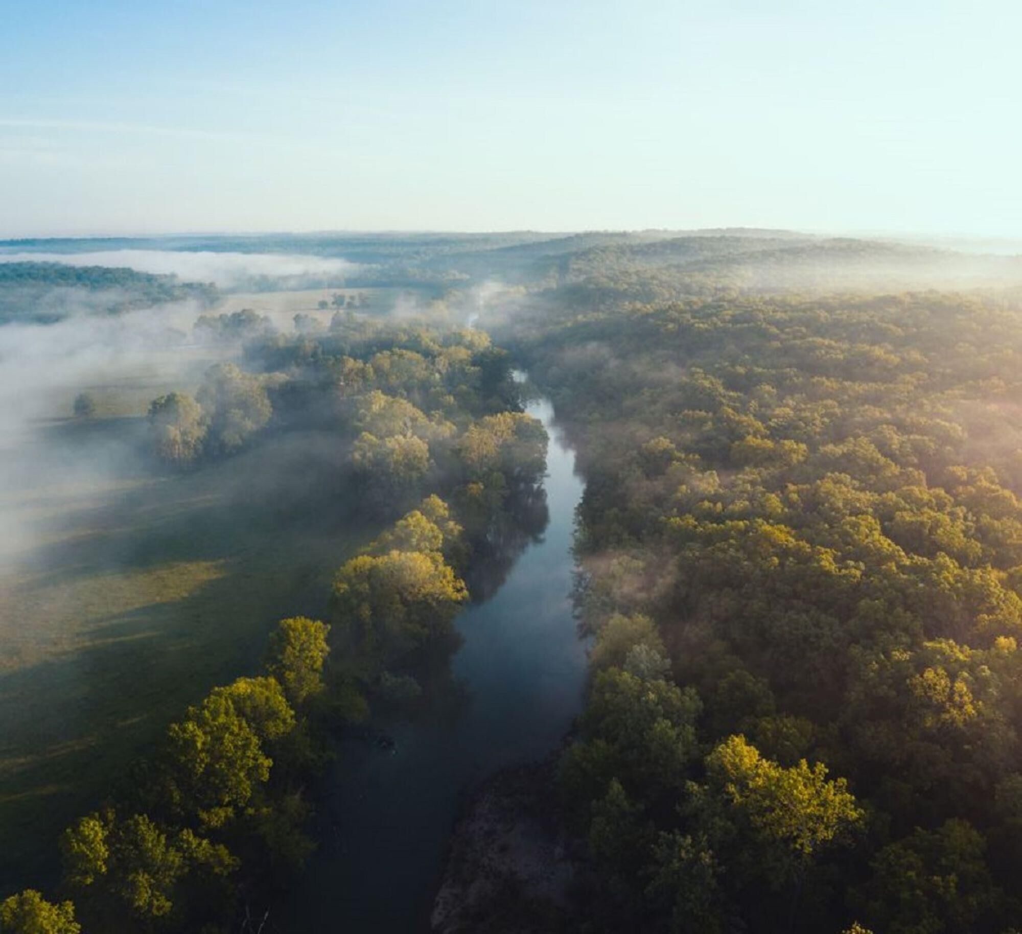Luchtbeeld van een rivier die zich een dicht bebost gebied slingert, met mist en heldere lucht.