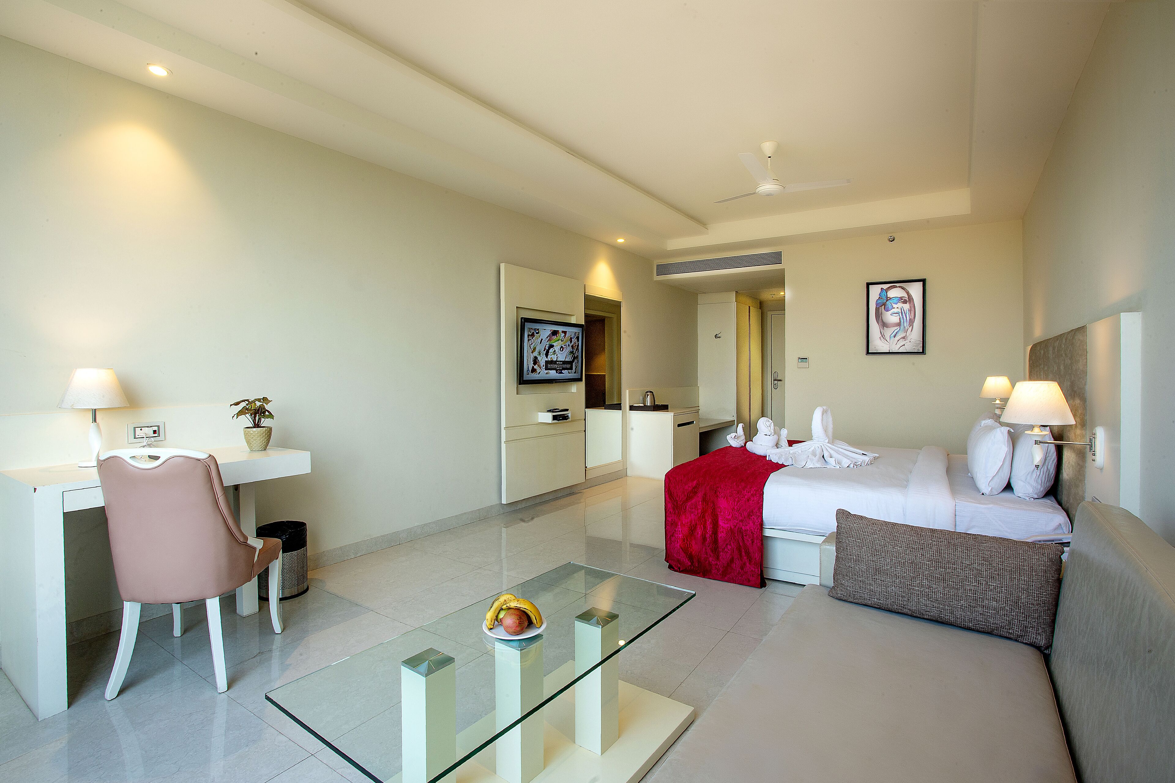 A hotel room with a bed, a desk, a chair, a glass coffee table, and a red throw blanket.