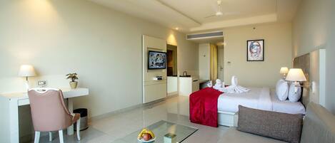 A hotel room with a bed, a desk, a chair, a glass coffee table, and a red throw blanket.