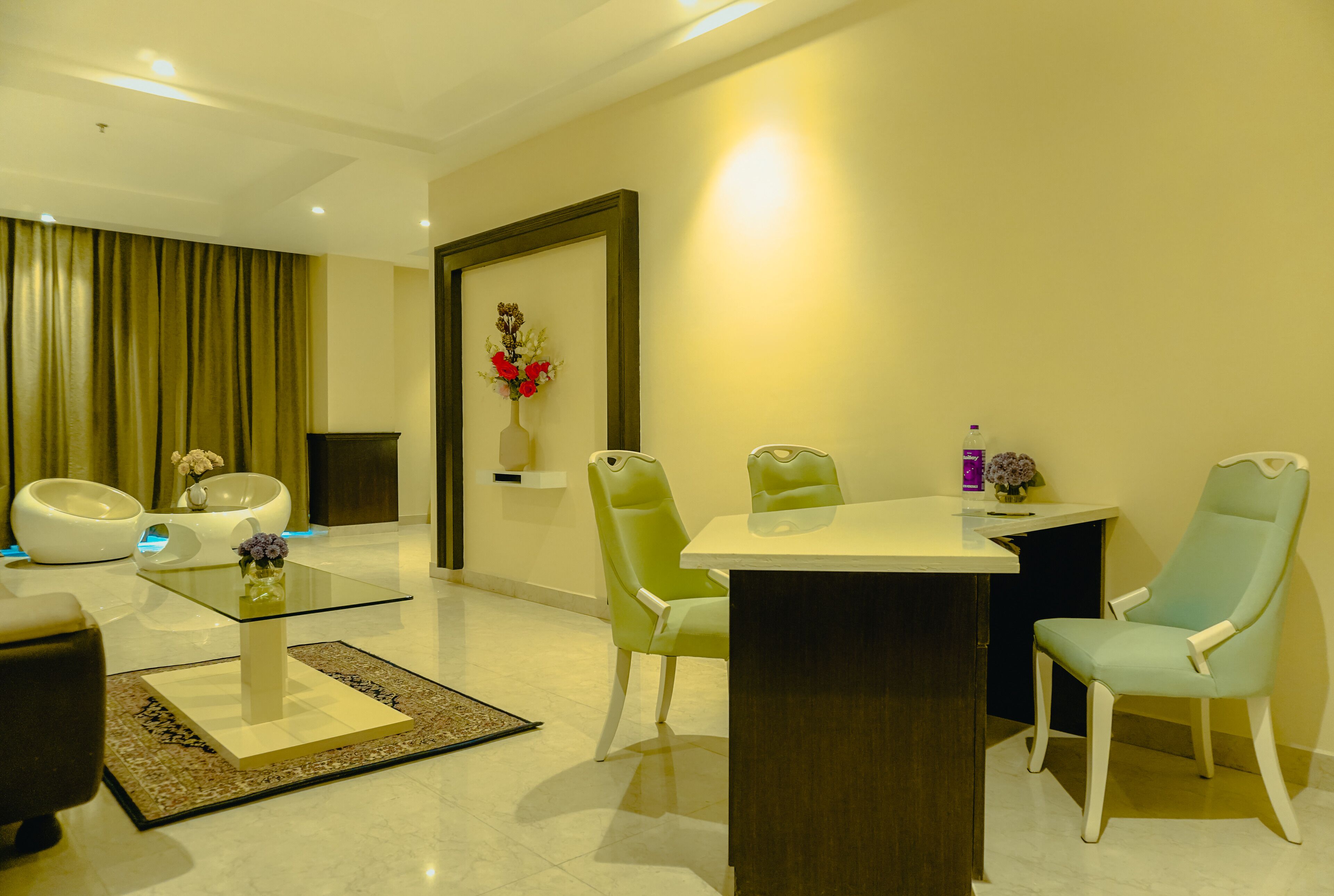 A modern dining area with a glass-top table, green chairs, and a white vase with flowers.
