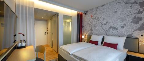 A modern hotel room with a large bed, a desk with a chair, a map on the wall, and a separate bathroom area.