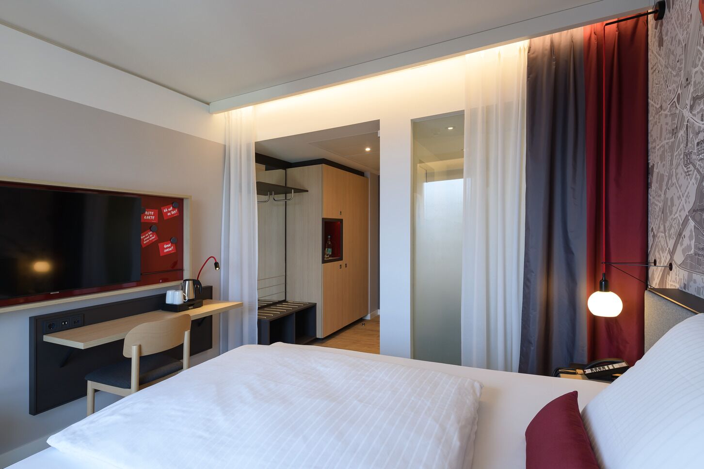 A modern hotel room with a bed, a desk, a TV, and a map on the wall.