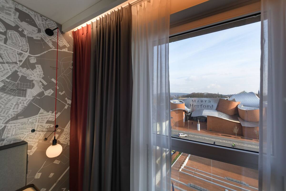 A hotel room with a large window, a city map on the wall, and a view of a modern building named "Marta Herford".