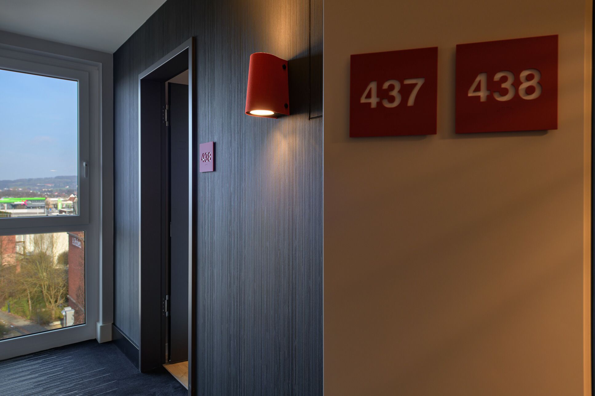 A hallway with a door leading to a room, a window with a view of buildings, and two red signs with numbers 437 and 438.