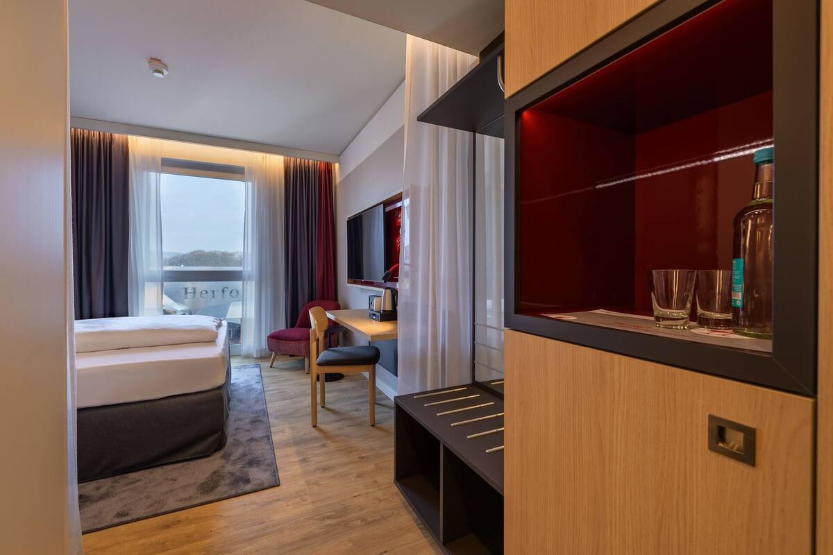 A modern hotel room with a bed, a desk, a chair, a TV, and a minibar.
