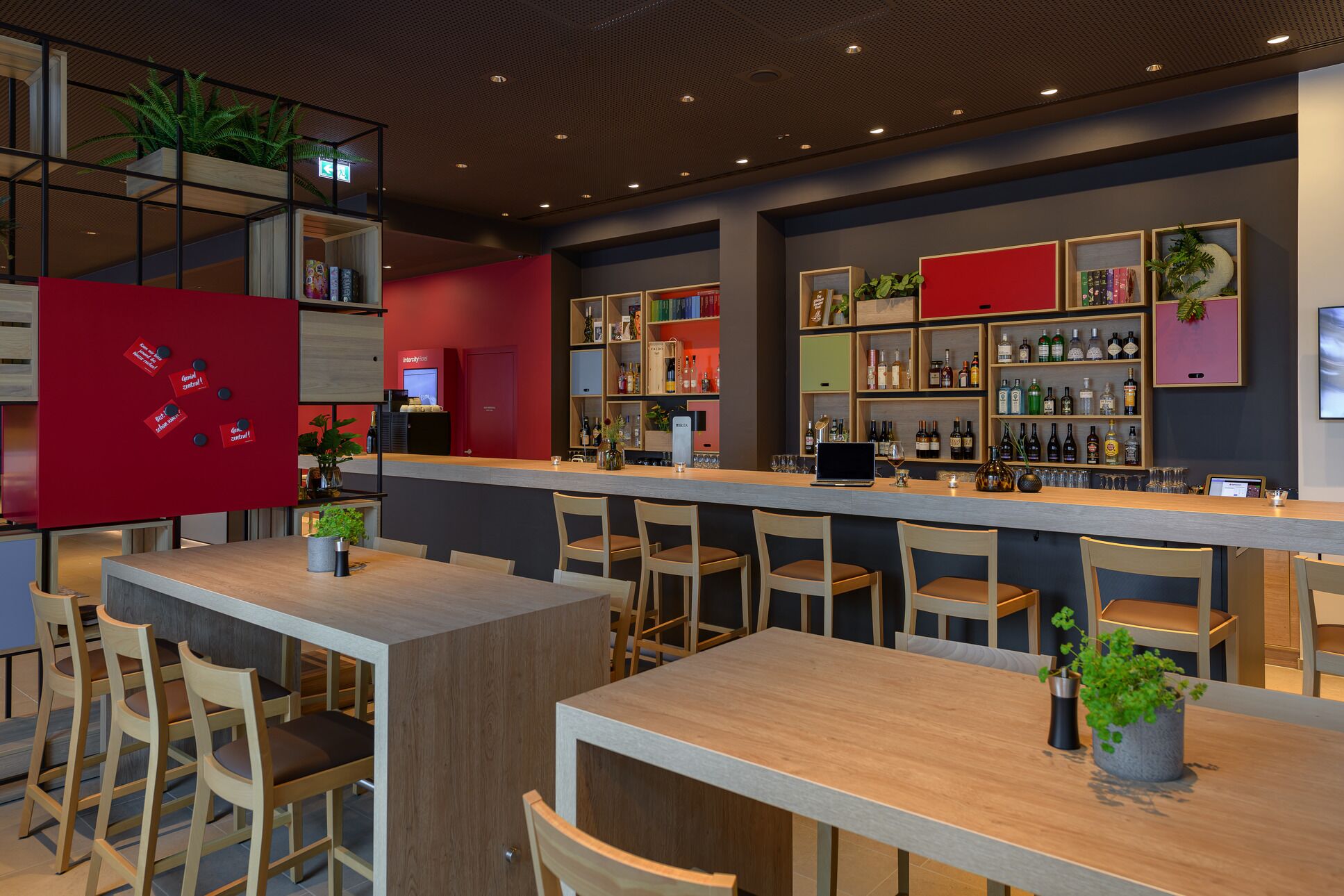 A modern bar area with wooden tables and chairs, shelves with bottles, and a red accent wall.