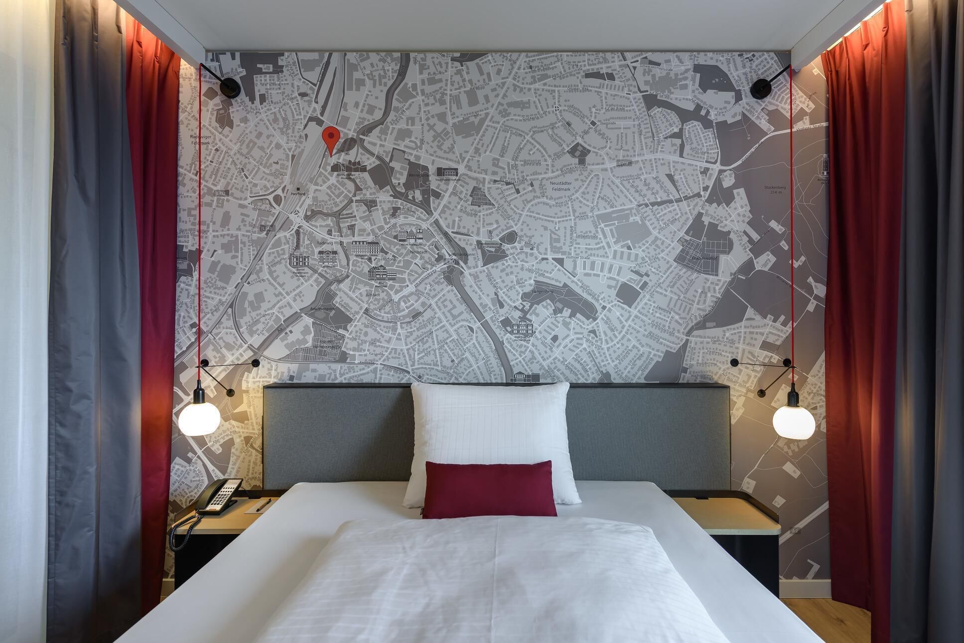 A hotel room with a large map on the wall, a bed with white and red bedding, two bedside tables with lamps, and red curtains.