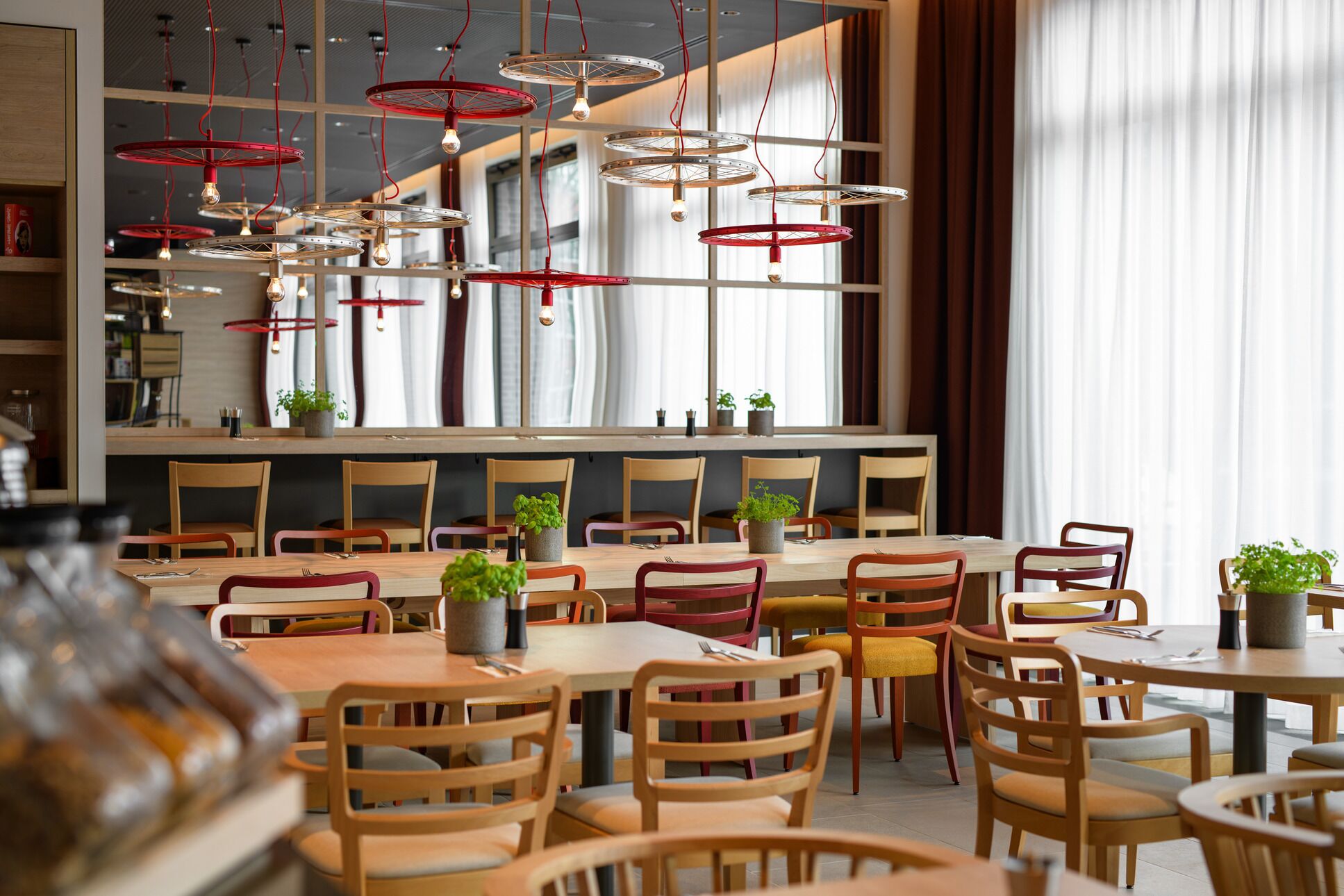 A modern restaurant interior with multiple dining tables and chairs, pendant lights, and large windows with curtains.