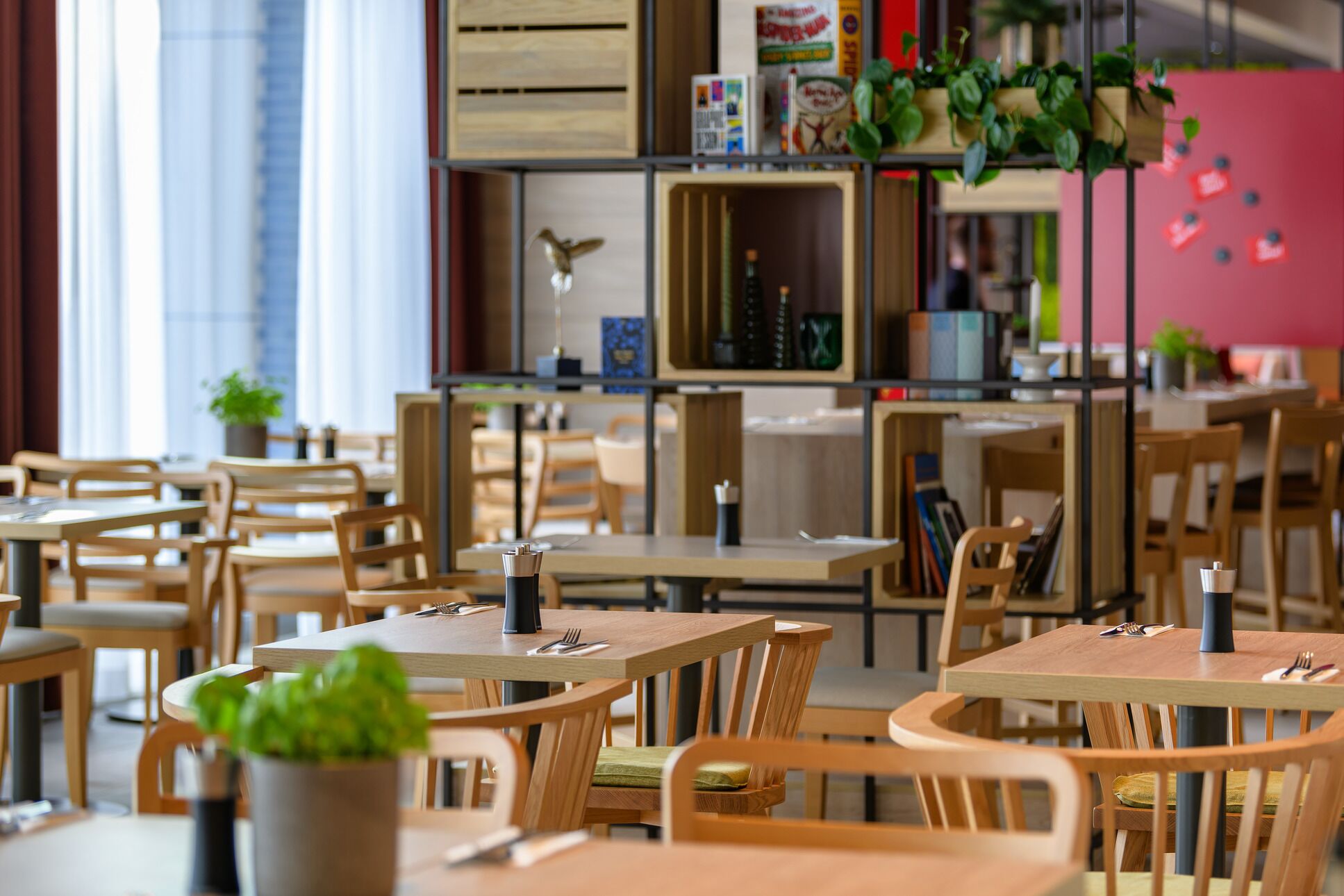 A modern dining area with wooden tables and chairs, some with plants. There are bookshelves with various items and a pink wall in the background.
