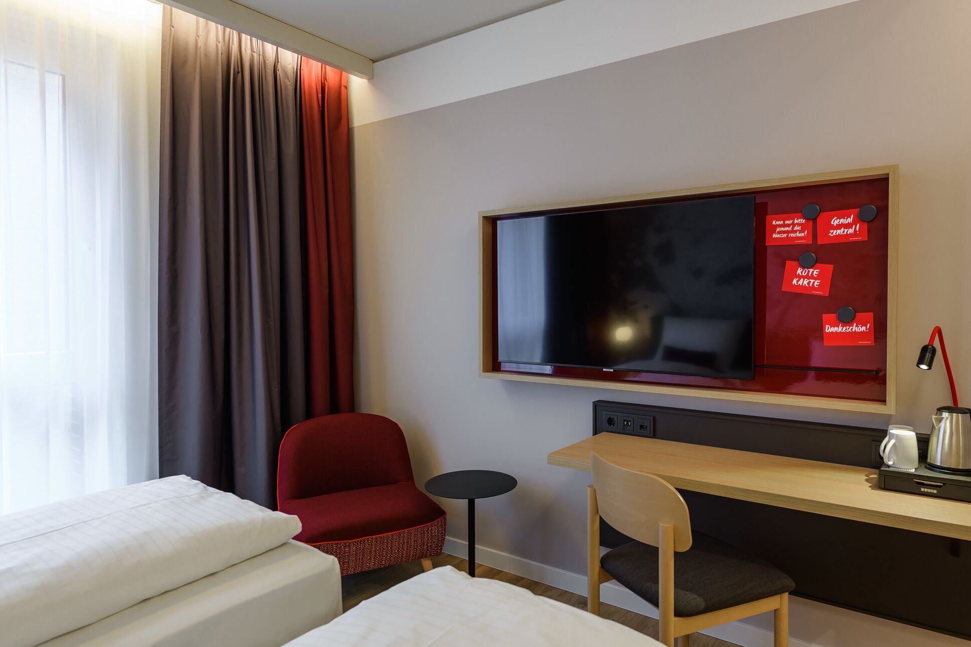 Habitación de hotel con cama, escritorio, silla, televisión y pared roja con letreros.