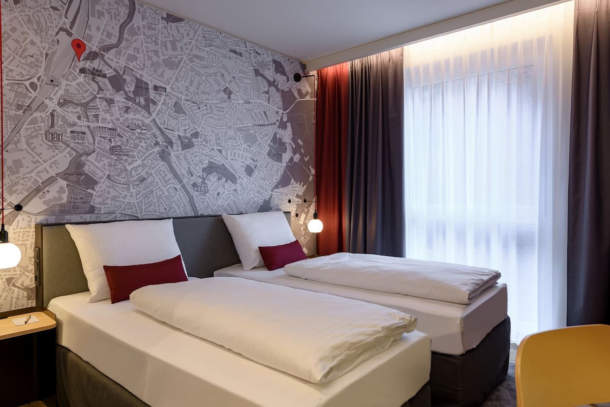 A hotel room with two beds, a map wall, a window with curtains, and a desk with a lamp.