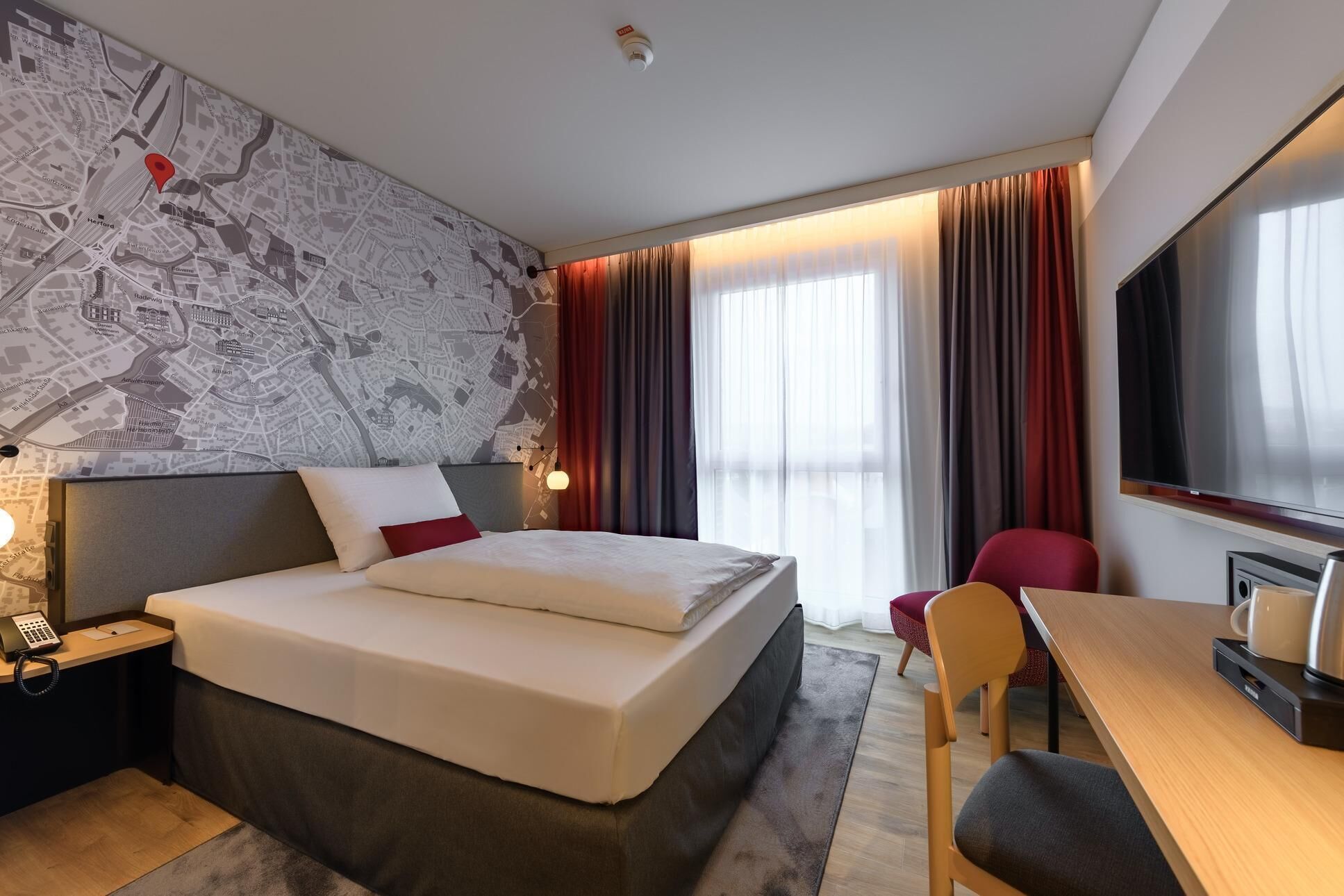 Une chambre d’hôtel avec un grand lit, un bureau, une chaise, une carte accrochée au mur et une fenêtre avec des rideaux.