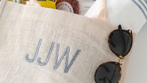 A woven tote bag with the initials "JJW" embroidered in blue, accompanied by a pair of sunglasses and a straw hat. - Charming Downtown 1 BR (Charleston)