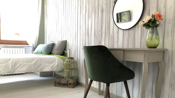 Un dormitorio con cama, una silla verde, una mesita de noche con un jarrón de flores y un espejo en la pared.