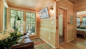 A cozy cabin interior with a wooden interior, a mounted sign for 'AYRES Vacation Rentals', a large window, a seating area with a leather couch, and a bed with a plaid bedspread. - Maple Treehouse | Unique, Private, Romantic (Gatlinburg)