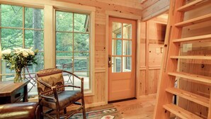 Interior de una cabaña de madera con un ventanal grande, una silla con cojín estampado y una estantería de madera.