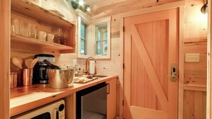 Cocina compacta con estantes de madera, una cafetera, un lavamanos y una puerta de diseño singular.