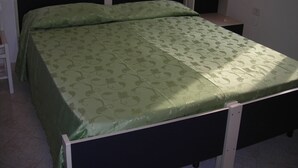 Un lit simple avec une couvre-lit à motifs verts, dans une pièce aux murs blancs et agrémentée d’une petite table de chevet avec une lampe.