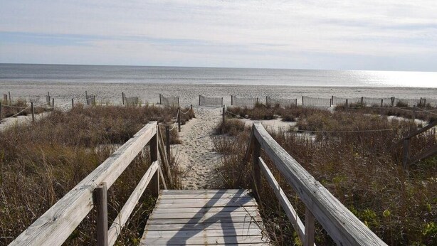 A wooden boardwalk leading to a sandy beach with a fence in the background.