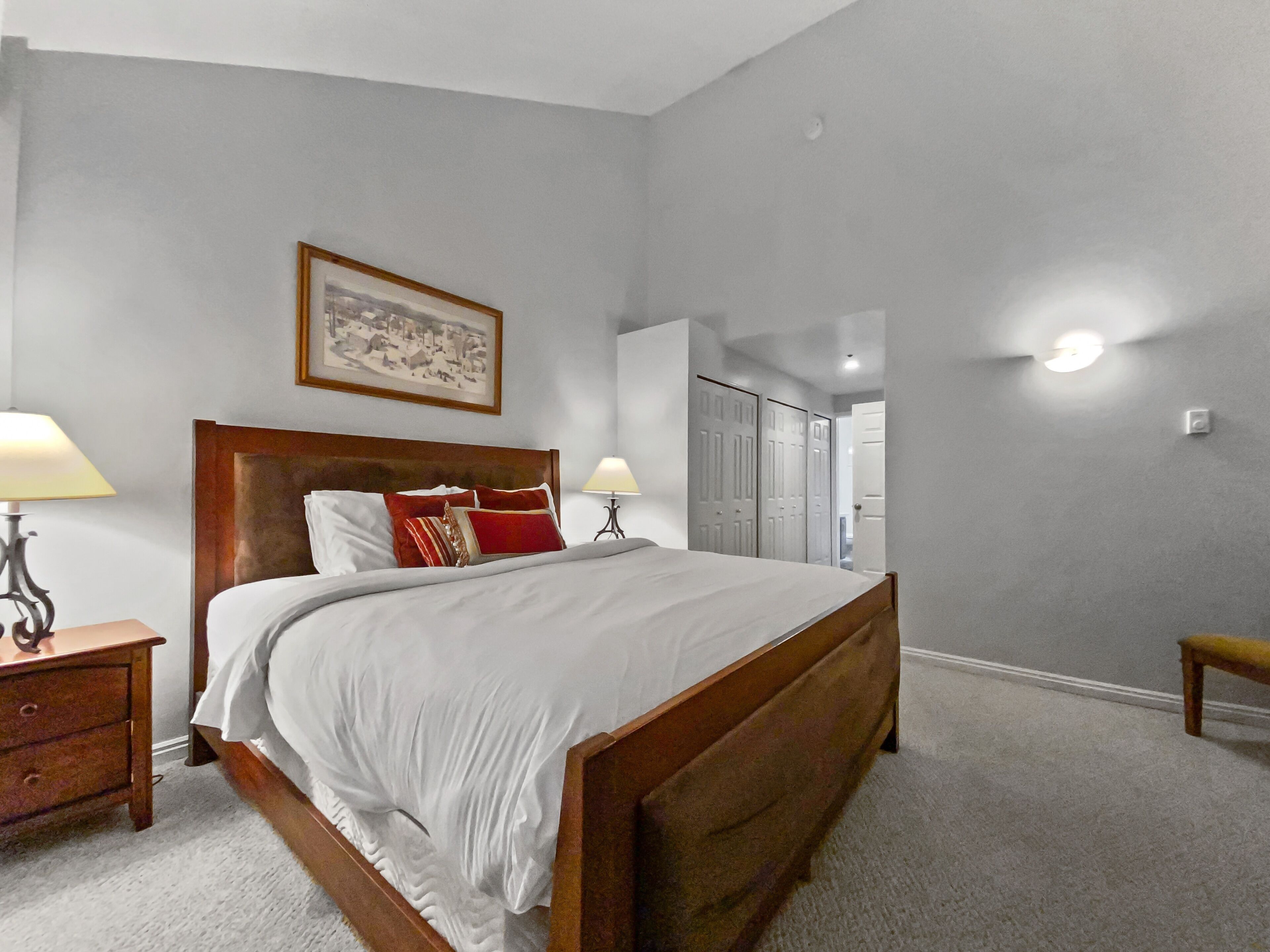 A bedroom with a wooden bed, two bedside tables, a nightstand, a framed picture on the wall, and a door leading to another room.