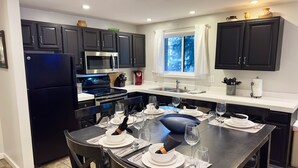 A modern kitchen with dark cabinets, a stainless steel refrigerator, a microwave, and a stove. There is a dining table set with plates, glasses, and cutlery.