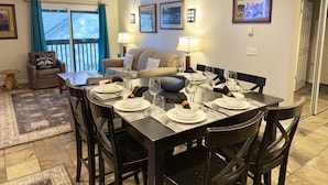 A dining area with a table set for six, a sofa, a chair with a pillow, and a window with curtains.