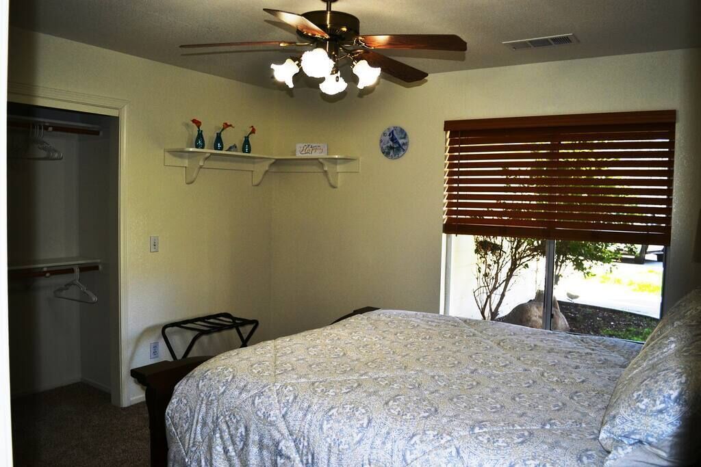 A bedroom with a bed, a ceiling fan, a window with blinds, a wall clock, and a small shelf with decorative items.